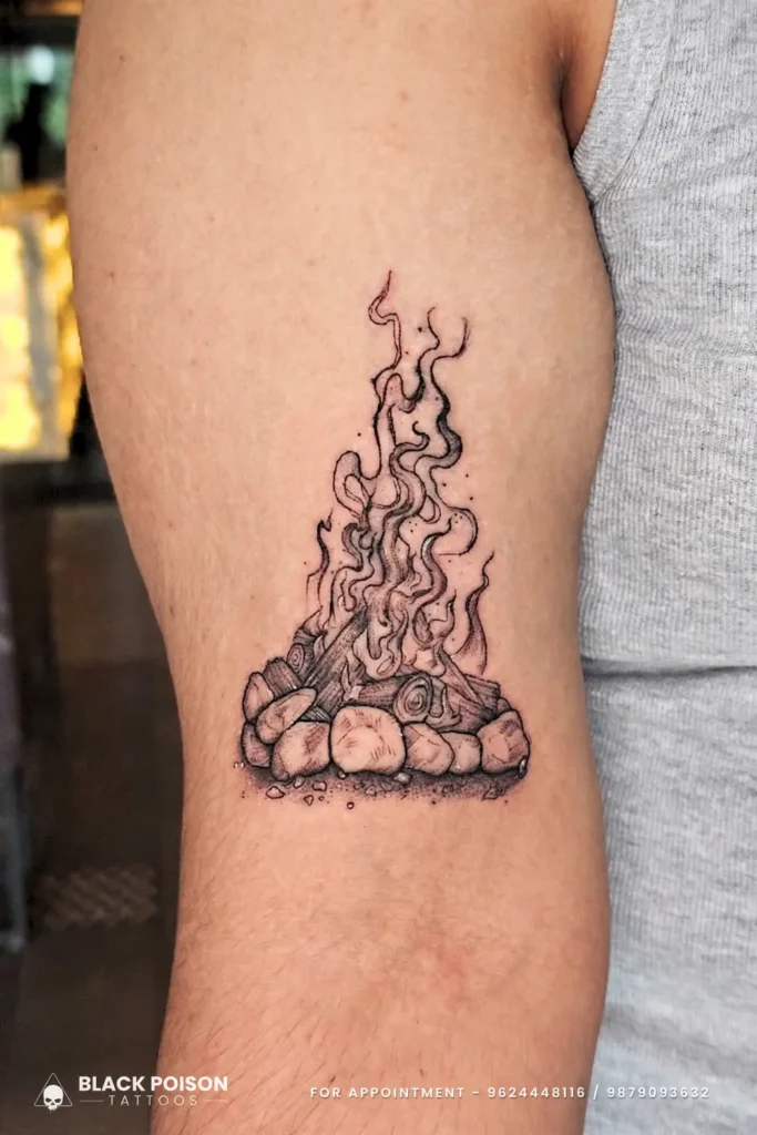 Fineline illustrative campfire tattoo with burning logs and rising smoke on upper arm by Black Poison Tattoo Studio Ahmedabad.