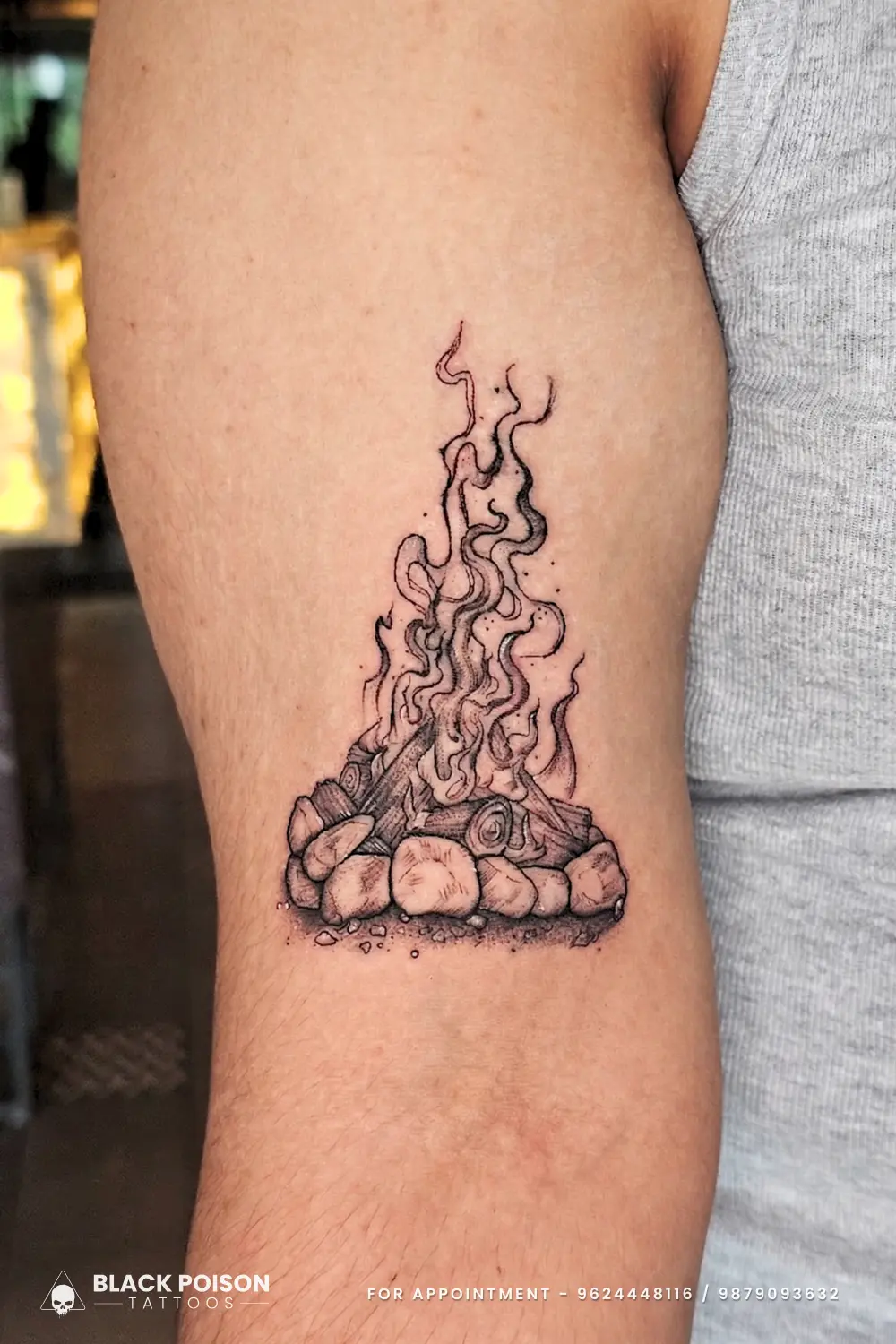 Fineline illustrative campfire tattoo with burning logs and rising smoke on upper arm by Black Poison Tattoo Studio Ahmedabad.
