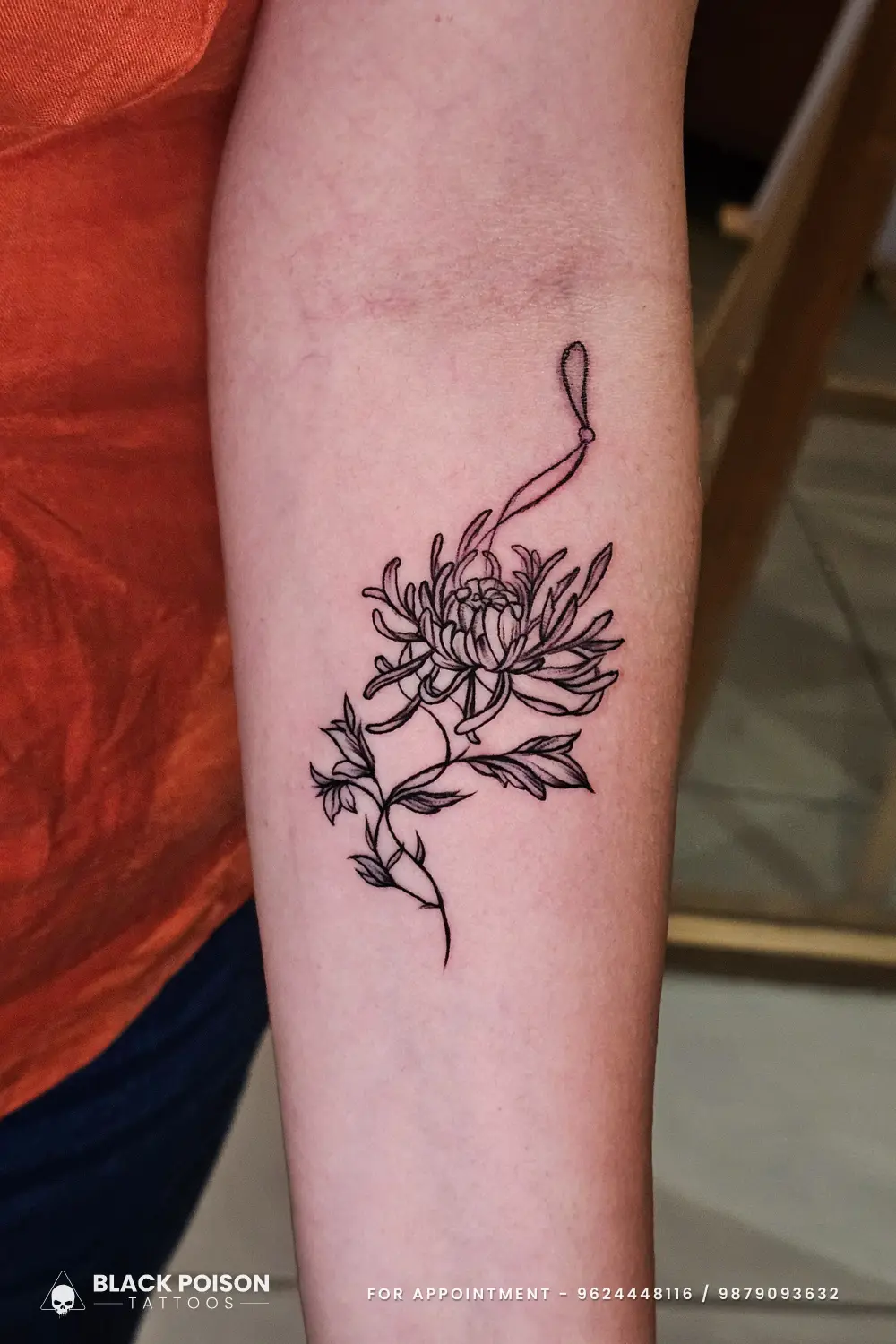 Delicate fine line chrysanthemum flower tattoo with a ribbon loop on the inner forearm, done at Black Poison Tattoos in Ahmedabad.