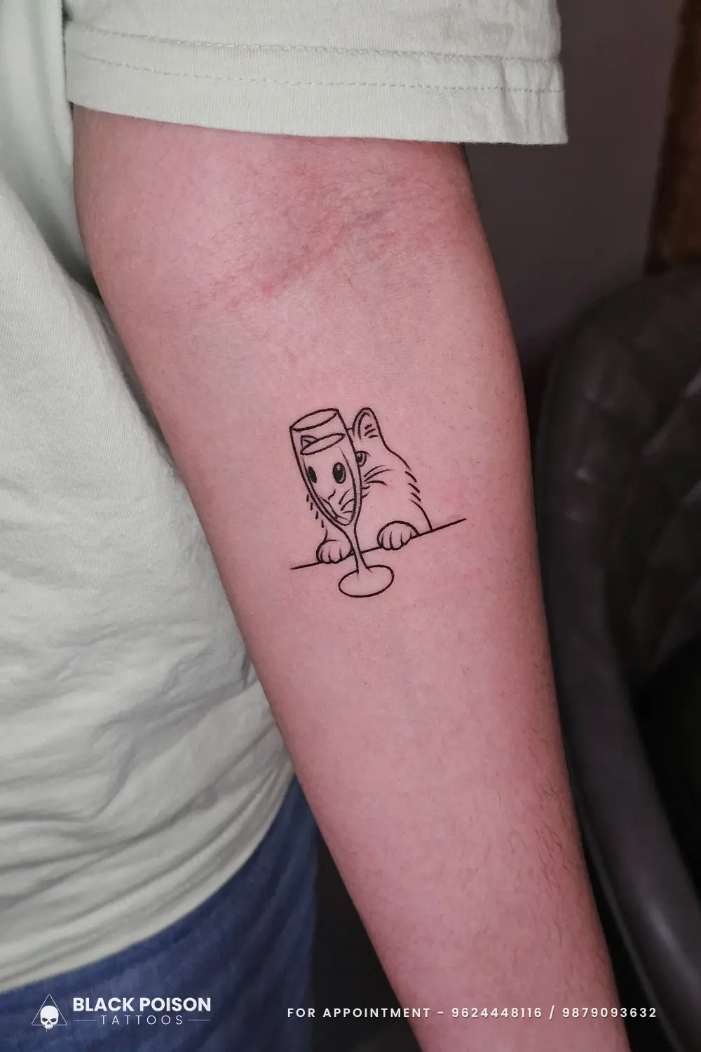 A minimalist fine line tattoo of a cat peeping through a wine glass, featuring distorted facial features for a humorous effect, inked on the inner forearm at Black Poison Tattoos Ahmedabad.