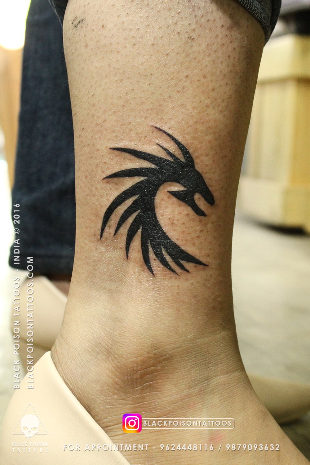 Bold black tribal dragon head tattoo with flowing spikes on the ankle by Black Poison Tattoo Studio Ahmedabad