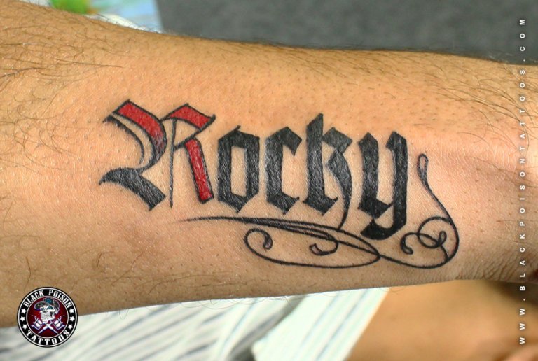 Name tattoo in bold gothic lettering with red shading on the forearm