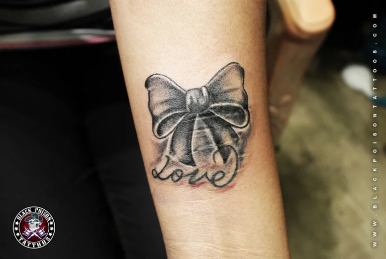 Black and grey ribbon bow tattoo with the word love on the forearm