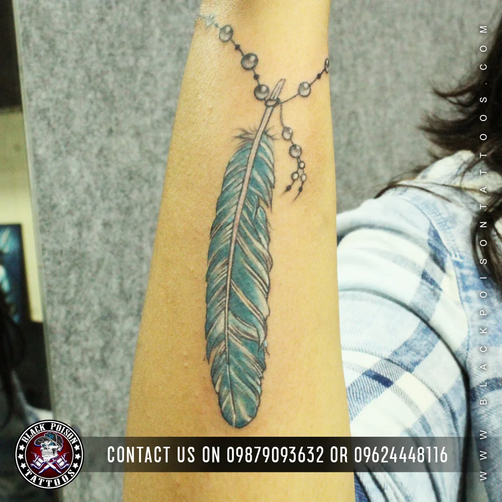 Teal and black feather tattoo with a hanging bead chain on the forearm