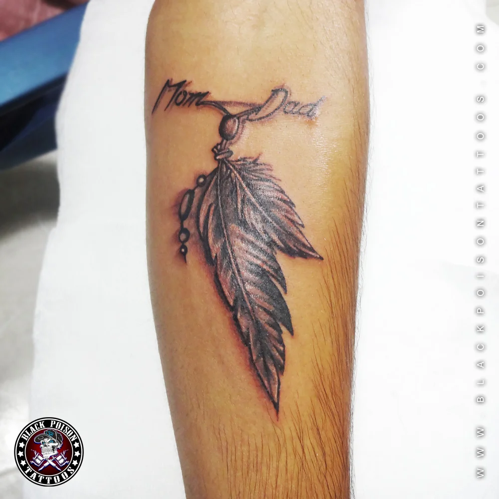 Mom and Dad script tattoo with two hanging feathers on the upper arm