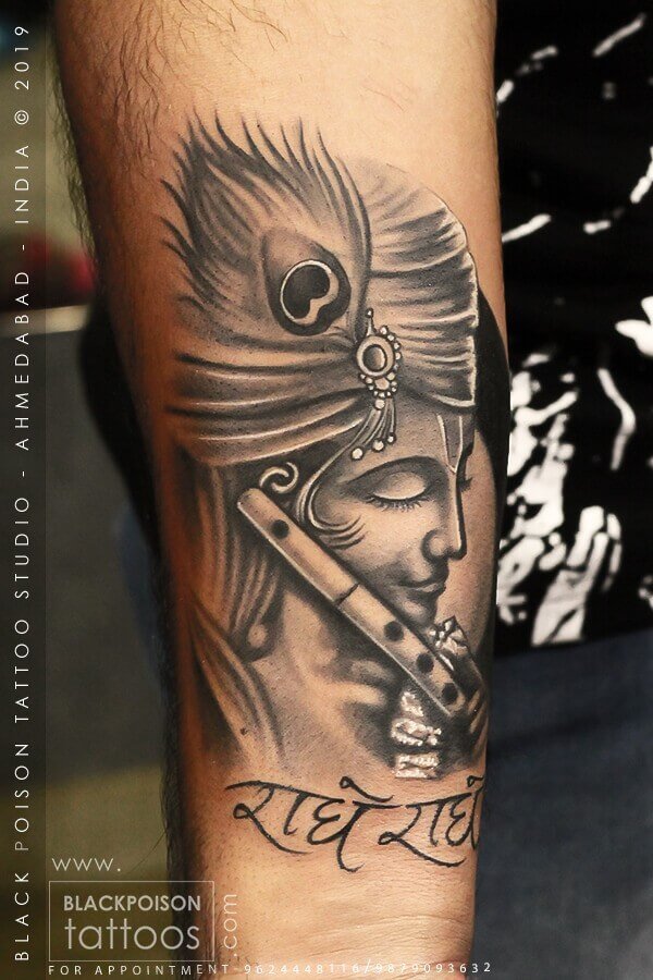 Black and grey Krishna tattoo with a flute, peacock feather and Radhe Radhe lettering on the forearm