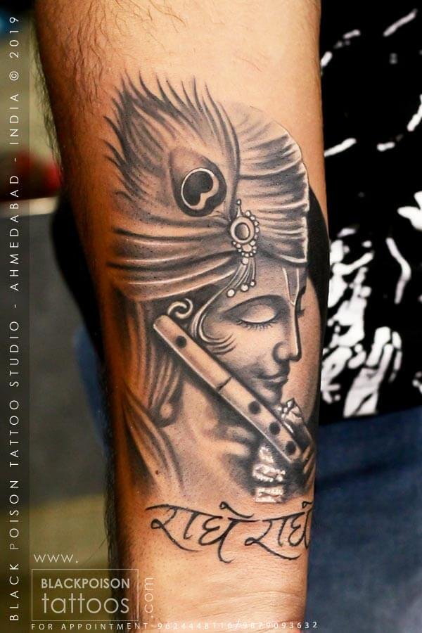 Black and grey Krishna tattoo with a flute and Radhe Radhe lettering on the forearm