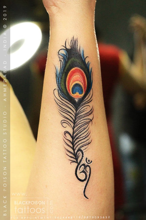Colourful peacock feather tattoo with a curled Om flourish on the forearm by Black Poison Tattoo Studio Ahmedabad