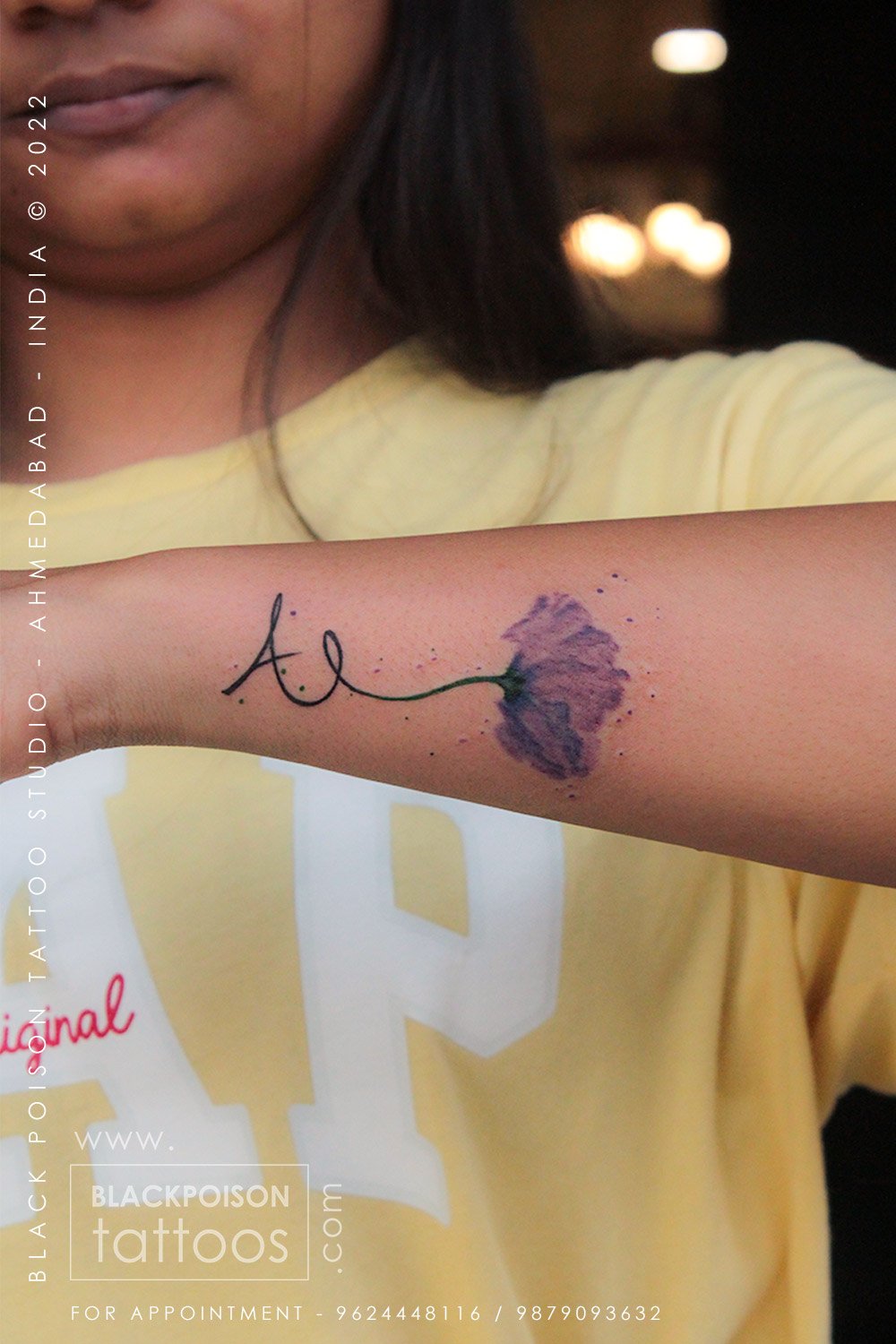 Fine line letter A tattoo joined to a purple watercolour flower on the forearm
