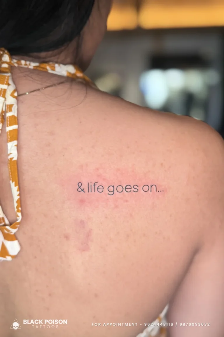Tiny black lettering tattoo reading and life goes on high on the shoulder blade at Black Poison Tattoo Studio, Ahmedabad