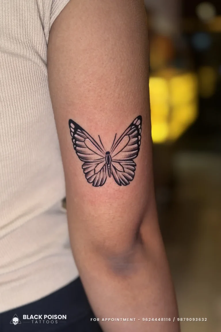 Black and grey butterfly with open wings and detailed veining on the outer upper arm at Black Poison Tattoo Studio, Ahmedabad
