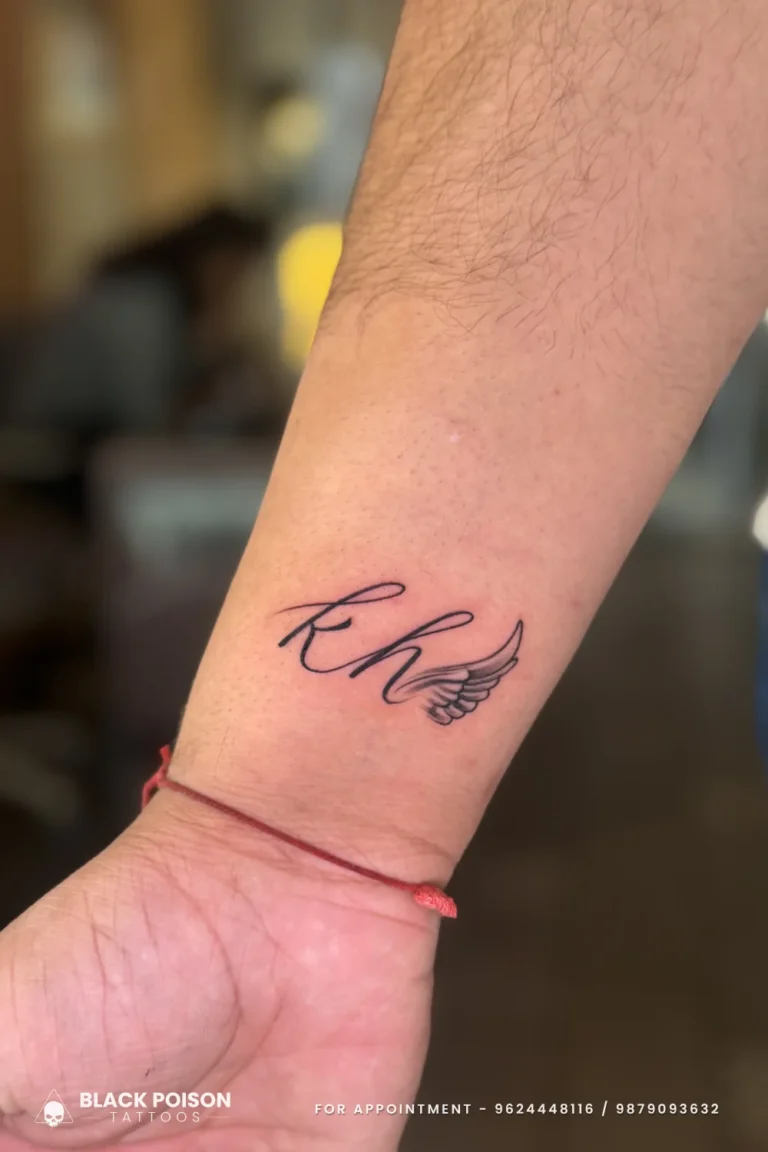 Cursive initials tattoo with a tiny angel wing in black on the inner wrist at Black Poison Tattoo Studio, Ahmedabad