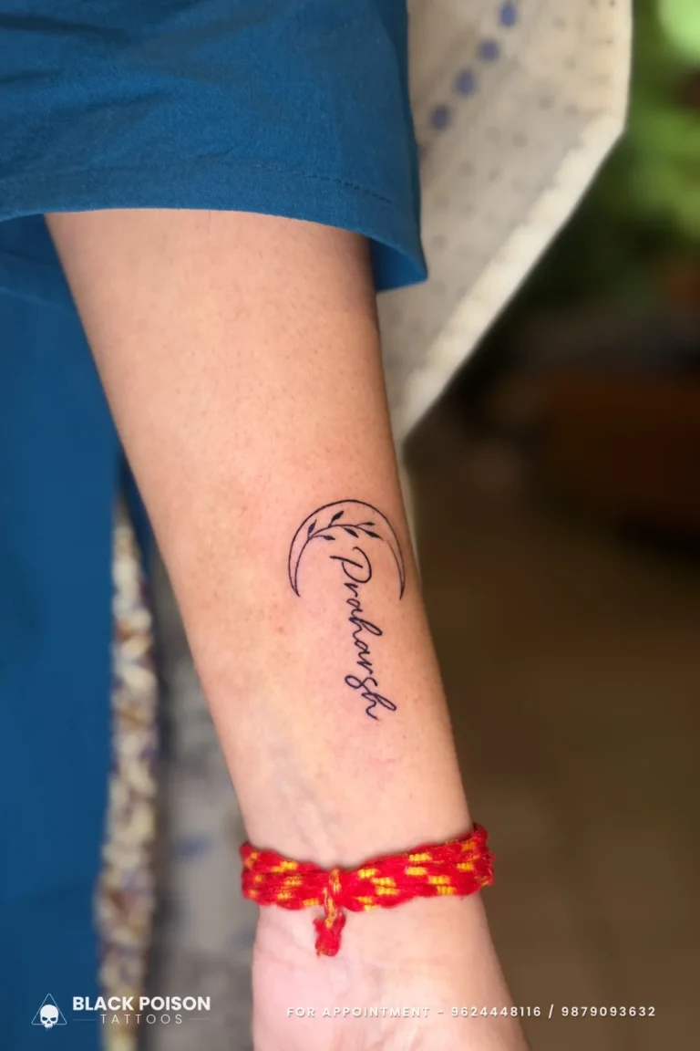 Cursive name tattoo in a crescent moon with a leaf sprig on the forearm at Black Poison Tattoo Studio, Ahmedabad