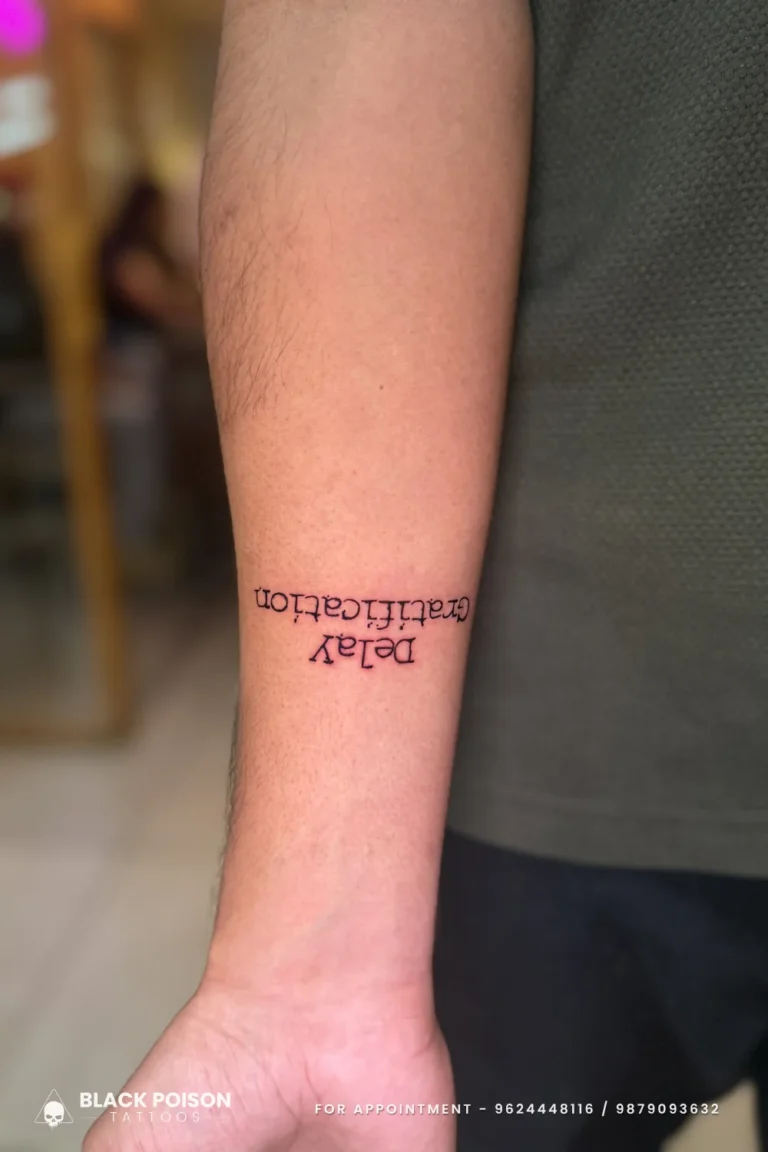 Black typewriter font tattoo reading Delay Gratification on the inner forearm at Black Poison Tattoo Studio, Ahmedabad