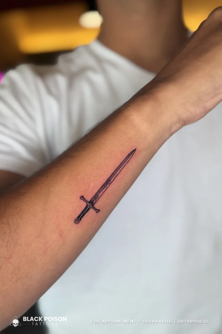 Slim fine line sword with a straight blade and crossguard along the outer forearm at Black Poison Tattoo Studio, Ahmedabad