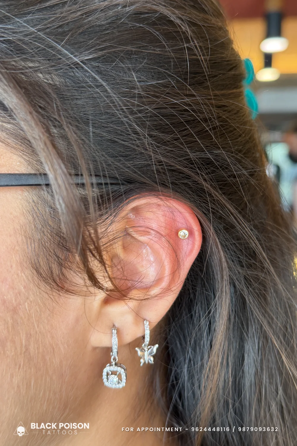 Flat piercing with a small gold crystal stud above a lobe wearing a square crystal drop hoop and a butterfly charm hoop