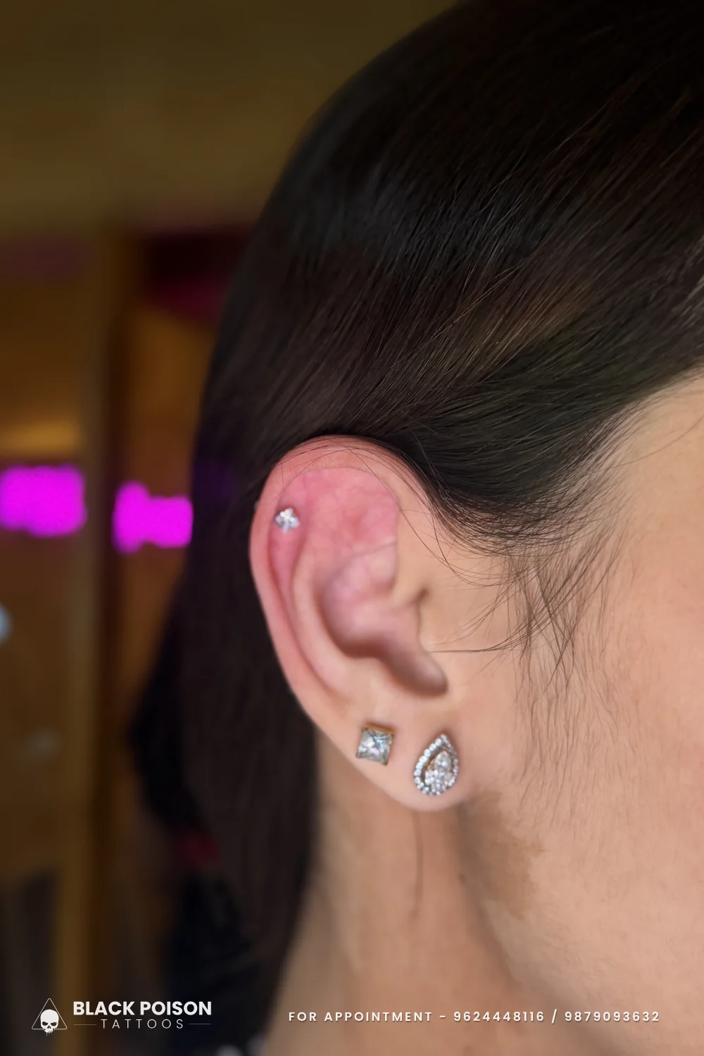 Freshly pierced helix with a tiny star stud, skin still pink, above a square crystal stud and a teardrop halo stud on the lobe