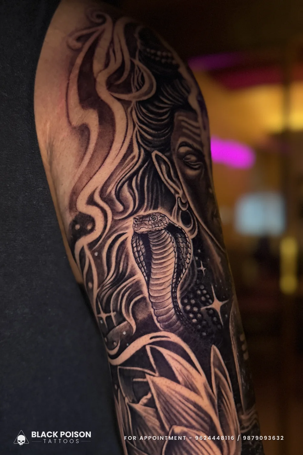 Black and grey Shiva face tattoo with a cobra rising in front of it and lotus petals below on the outer upper arm