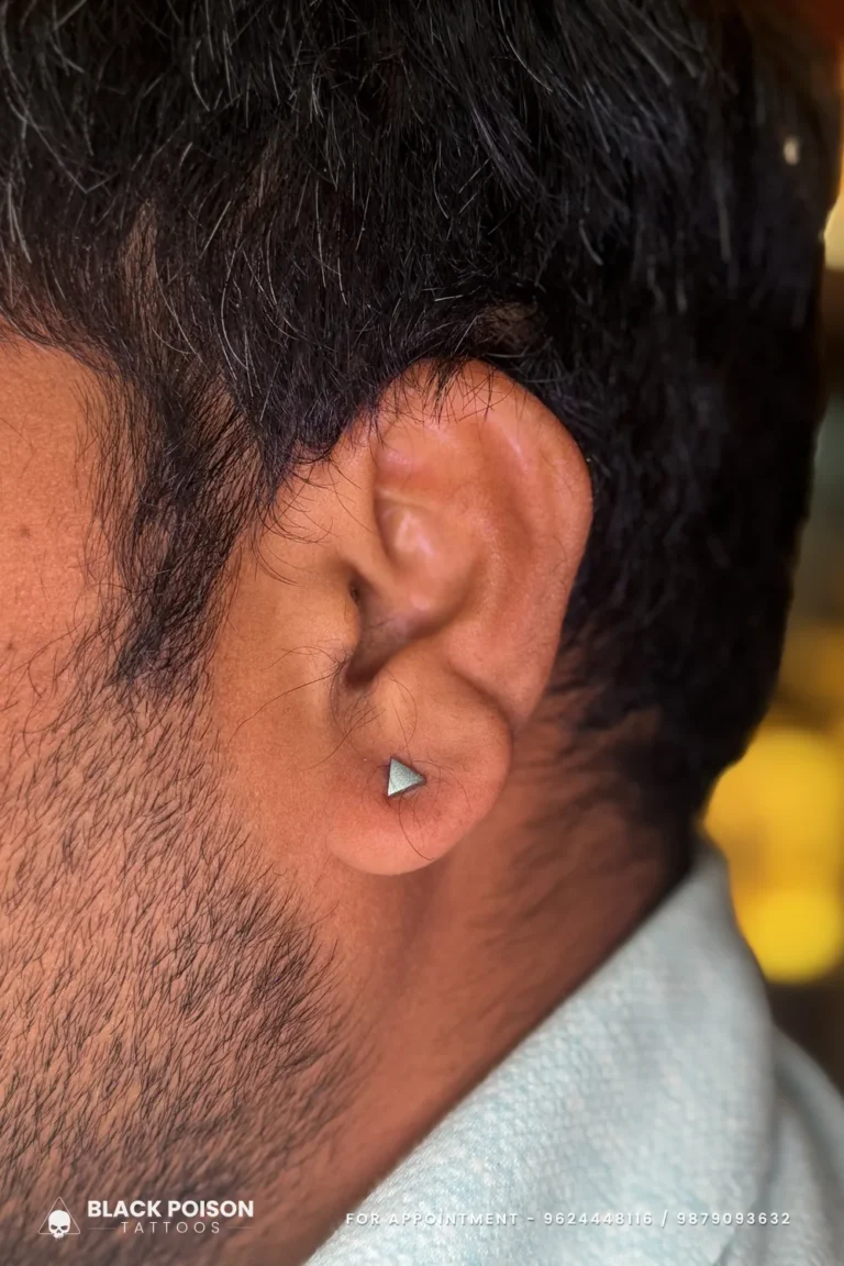 Men's ear lobe piercing with a small silver triangle stud, a minimal geometric look at Black Poison Tattoo Studio, Ahmedabad