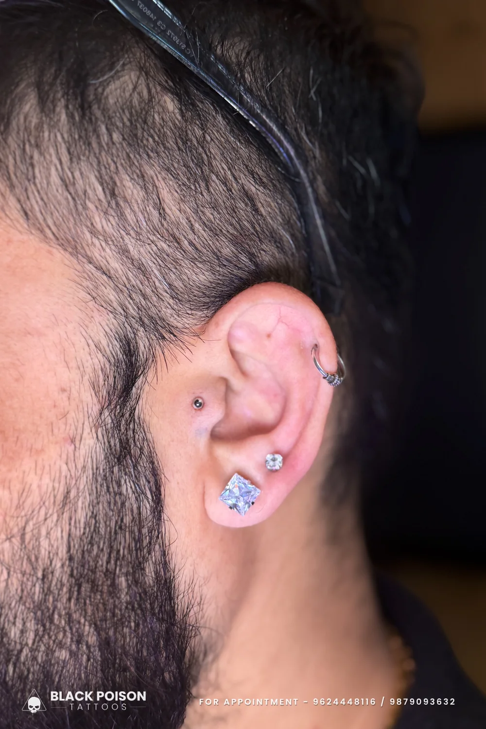Close-up of a men's ear with a matte black tragus stud, a slim helix ring and two crystal studs on the lobe