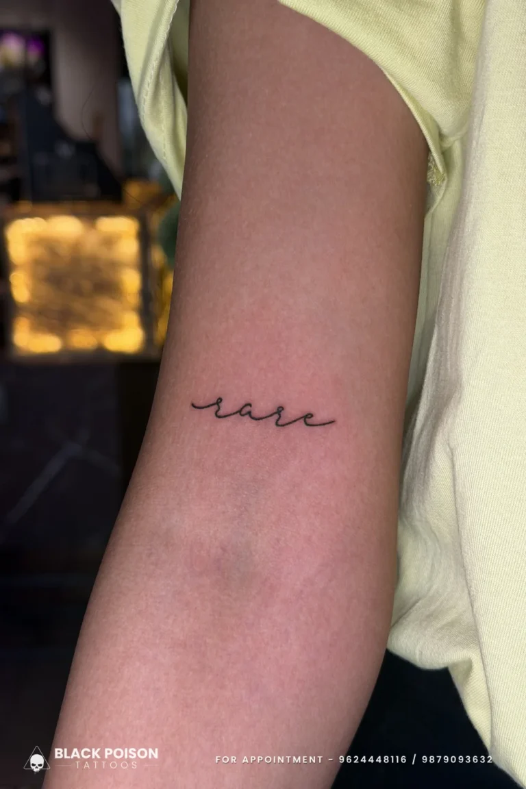 The word rare in delicate thin black cursive on the inner upper arm at Black Poison Tattoo Studio, Ahmedabad