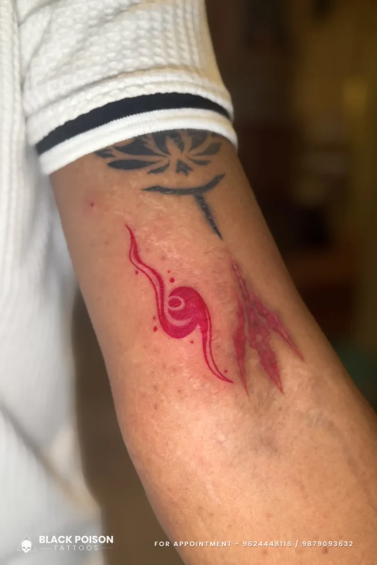 Red ink abstract trishul with a damru swirl and splash dots on the upper arm at Black Poison Tattoo Studio, Ahmedabad