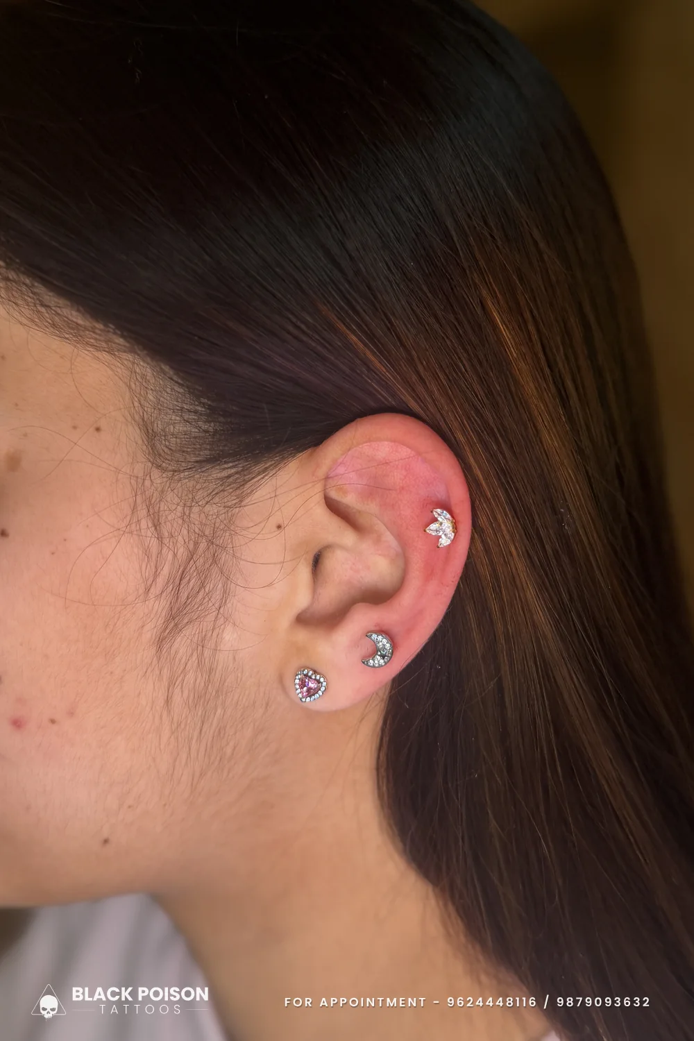 Stacked lobe piercings with a jewelled pink heart stud and a crescent moon stud, plus a leaf-cluster stud on the helix