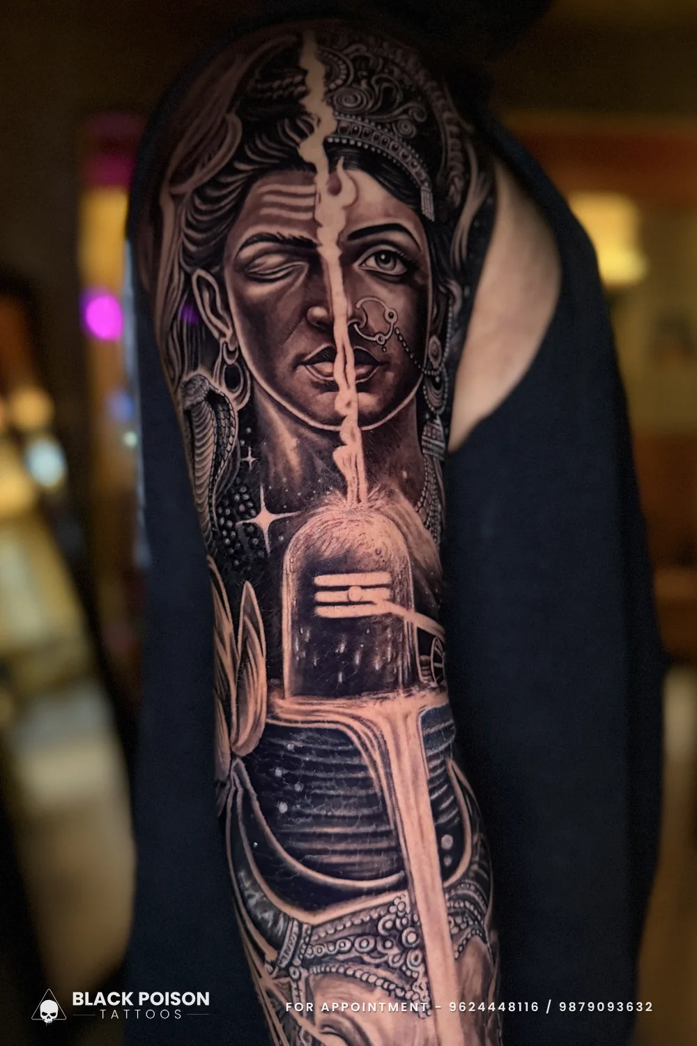 Black and grey Shiva Parvati split face tattoo on the upper arm flowing into the top of the Shivling below the elbow