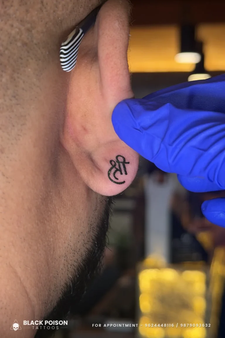 Bold black Devanagari Shree tattooed on the ear lobe, held by a gloved hand, at Black Poison Tattoo Studio, Ahmedabad