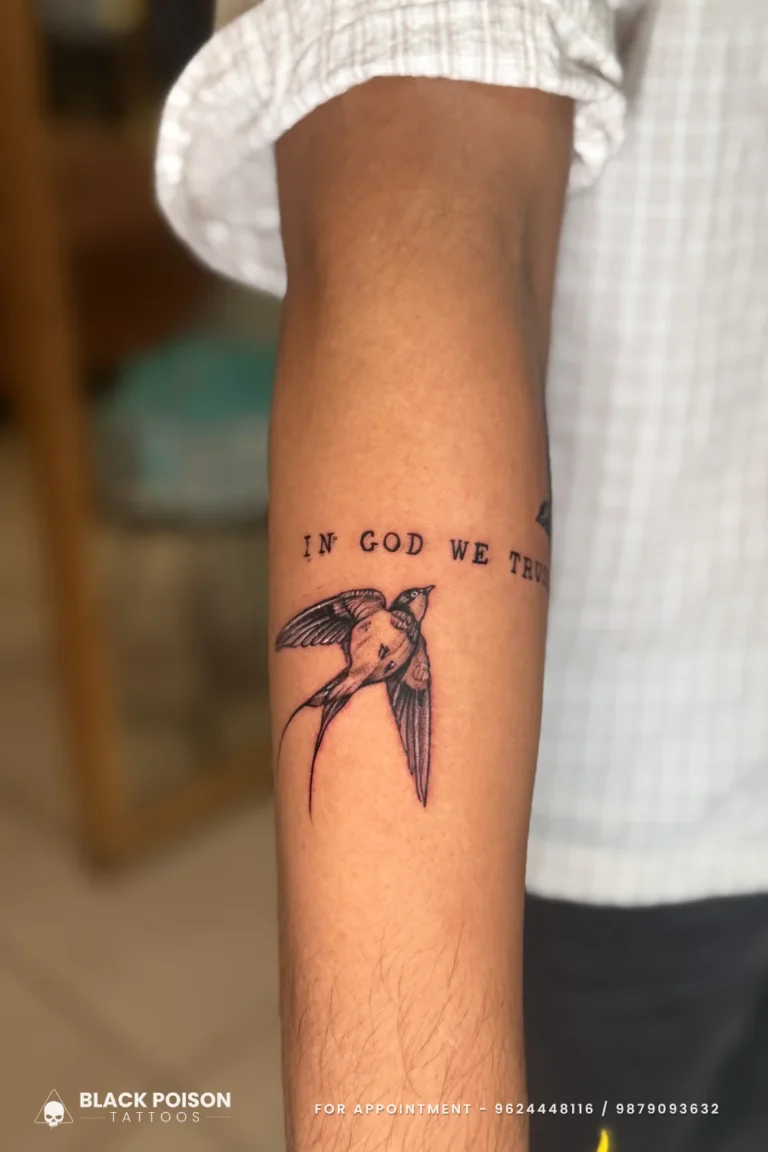 Black and grey swallow beneath In God We Trust lettering wrapping the forearm at Black Poison Tattoo Studio, Ahmedabad